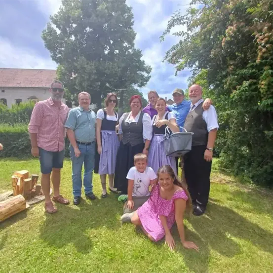 Eine Gruppe von Menschen steht auf einem Rasen und eine Frau sitzt vorne. Ein Mann hält eine Tasche. Sie scheinen für ein Foto zu posieren. Bäume und Büsche stehen hinter ihnen und ein Gebäude ist in der Ferne.