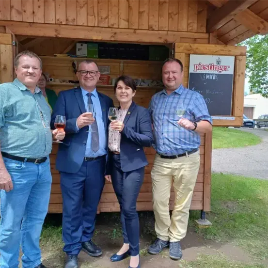 Vier Personen in formeller Kleidung stehen vor einer Holzhütte. Sie halten Weingläser und lächeln. Hinter ihnen steht ein Schild mit der Aufschrift 'Piestinger'.