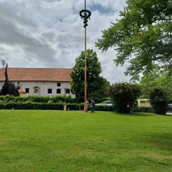 Zwei Personen stehen neben einem hohen Pfahl mit einem Kranz. Sie befinden sich auf einer Wiese mit einem Haus im Hintergrund.