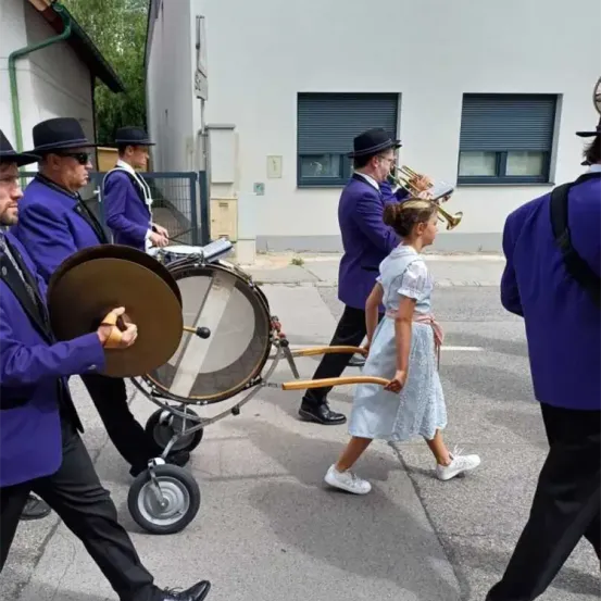 Eine Musikkapelle zieht die Straße entlang. Sie tragen lila Uniformen und Hüte. Ein Mann hält eine Trommel und ein Becken. Ein Mädchen geht vor ihnen. Dahinter steht ein Gebäude mit einem Tor und einem Baum.