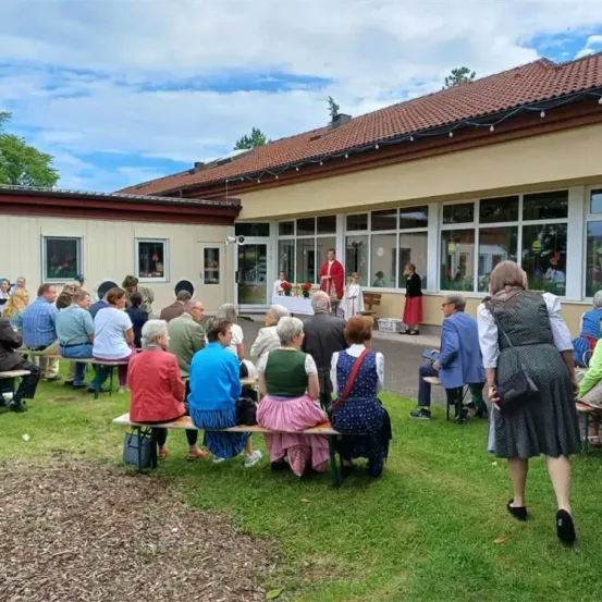 Eine Gruppe von Menschen sitzt auf Bänken auf einem Rasen vor einem Gebäude. Einige sind in traditioneller Kleidung gekleidet. Ein Mann und eine Frau stehen in der Nähe des Eingangs.