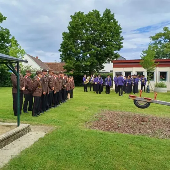 Eine Gruppe von Menschen in braunen Uniformen steht in einer Reihe auf einem Rasen. Dahinter spielt eine weitere Gruppe Blasinstrumente. Im Hintergrund befinden sich Bäume und Gebäude.
