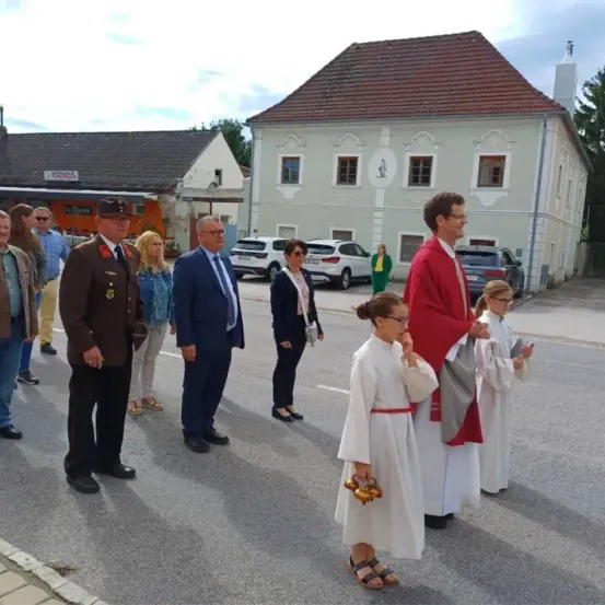 Eine Gruppe von Menschen steht auf einem Stadtplatz, einige in formeller Kleidung, andere in weißen Roben. Ein Gebäude mit rotem Dach steht hinter ihnen, und Autos sind davor geparkt.