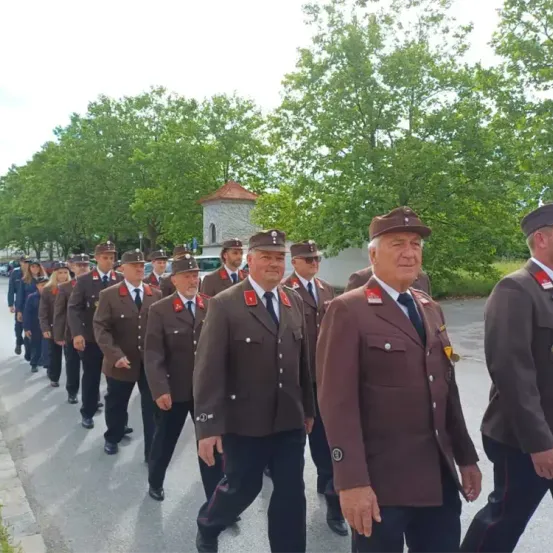 Eine Gruppe uniformierter Personen, wahrscheinlich Militär, marschiert bei einer Parade an einem sonnigen Tag mit Bäumen und Gebäuden im Hintergrund.