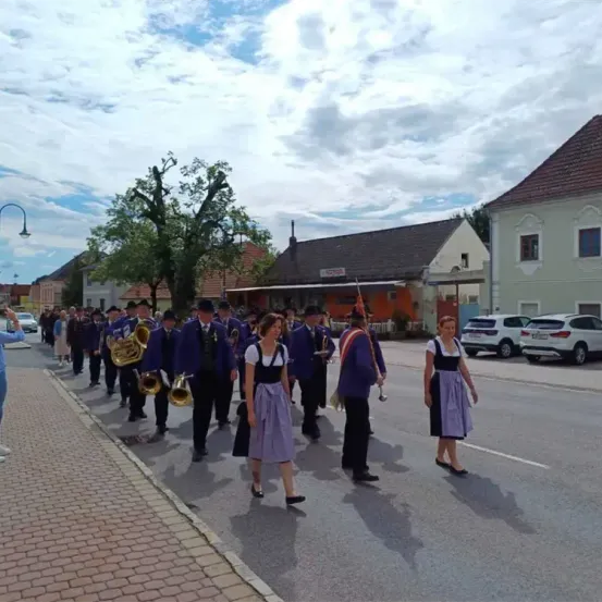 Eine Musikkapelle spielt auf der Straße, wobei Musiker verschiedene Instrumente spielen. Zuschauer beobachten von der Straße aus. Im Hintergrund sind geparkte Autos und Gebäude mit einigen Bäumen.