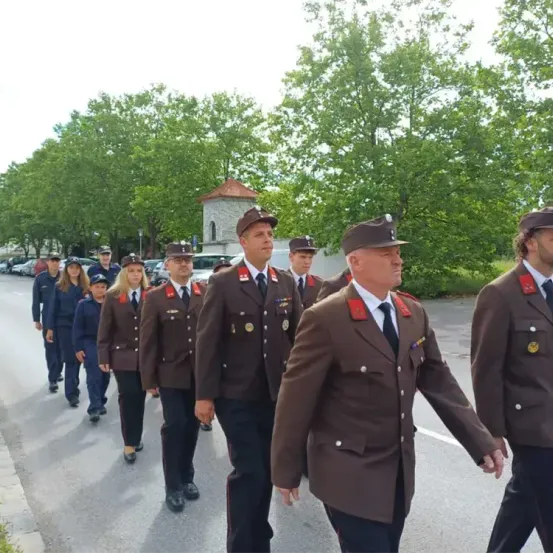 Eine Gruppe uniformierter Personen, möglicherweise Militärangehörige, marschiert eine Straße hinunter, gesäumt von Bäumen und geparkten Autos.