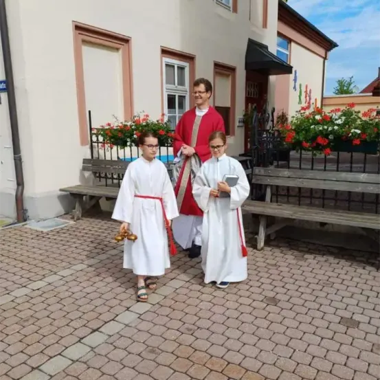 Ein Priester mit zwei jungen Kindern in weißen Gewändern geht vor einer Kirche.