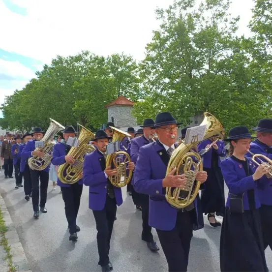 Ein Blasorchester in lila Uniformen spielt Blechblasinstrumente an einem sonnigen Tag, mit Bäumen und einem Gebäude im Hintergrund.