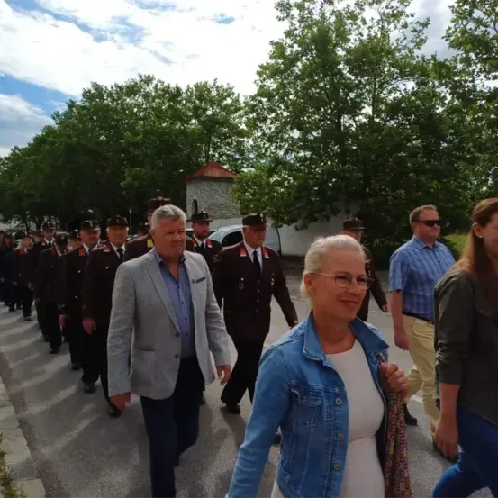 Ein Mann in Anzug und eine Frau in einer Jeansjacke gehen vor einer marschierenden Militärparade an einem sonnigen Tag.