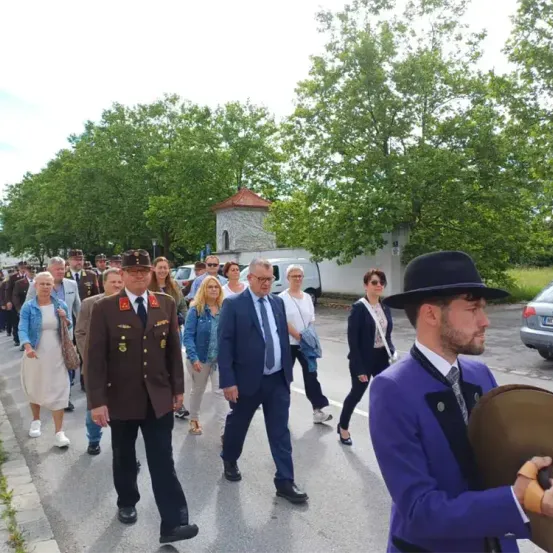 Eine Gruppe von Menschen geht auf der Straße, wahrscheinlich für eine Parade. Ein Mann in Uniform und ein Mann mit Hut führen die Gruppe an. Bäume und Gebäude säumen die Straße.