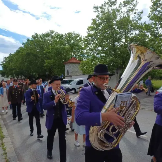 Eine Brassband marschiert an einem sonnigen Tag bei einer Parade, mit Musikern, die verschiedene Instrumente spielen, und Zuschauern, die vom Bürgersteig aus zuschauen.