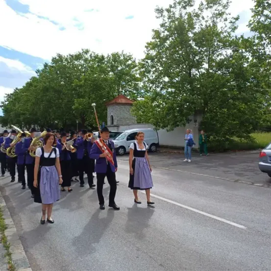 Eine Blaskapelle spielt auf der Straße mit mehreren Leuten, die Blechblasinstrumente spielen. Eine Person mit einem Taktstock führt die Band an. Einige Leute stehen am Straßenrand und beobachten sie.