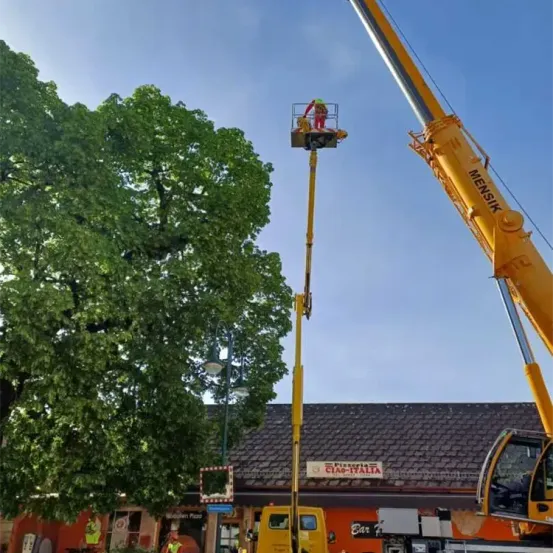 Eine Person mit grünem Helm steht in einem Korblift, der an einen hohen gelben Kran angeschlossen ist, vor einem Gebäude mit einem Schild, das Pizzeria Ciao-Italia liest.