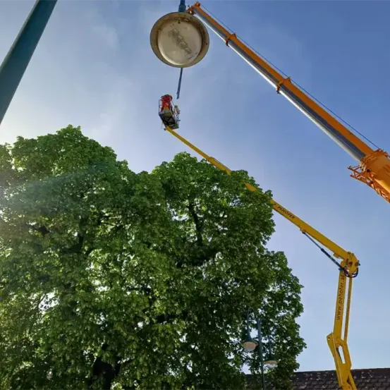 Eine Person in einer Arbeitsbühne erreicht eine Straßenlaterne. Die Arbeitsbühne ist gelb. Der Himmel ist blau.