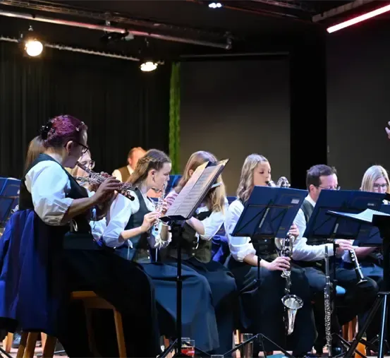 Eine Band spielt auf der Bühne mit Musikinstrumenten. Der Dirigent steht davor. Die Musiker sitzen auf Stühlen mit Notenständern davor. Einige Leute stehen hinter ihnen. Scheinwerfer sind an der Decke montiert.