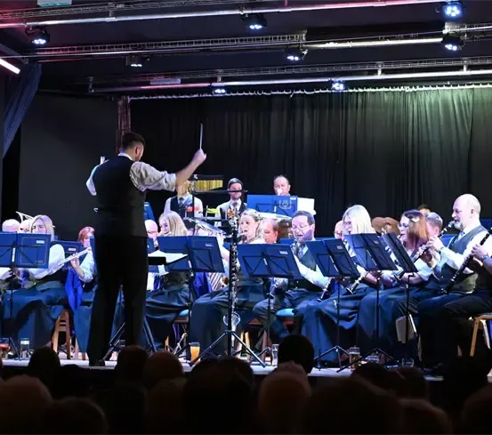 Ein Orchester spielt auf einer Bühne vor einem Publikum. Der Dirigent führt das Orchester mit einem Taktstock. Die Musiker sitzen auf Stühlen und spielen ihre Instrumente. Das Publikum beobachtet die Aufführung. Die Bühne ist mit Lichtern beleuchtet.