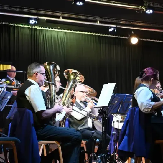 Ein Orchester spielt auf der Bühne mit verschiedenen Blech- und Holzblasinstrumenten unter Scheinwerfern. Musiker sitzen auf Stühlen mit Notenständern davor.