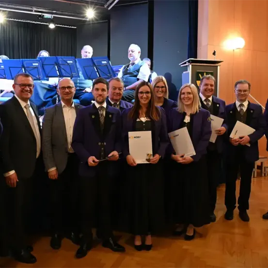 Eine Gruppe von formell gekleideten Personen steht zusammen und lächelt für die Kamera. Sie halten Urkunden in den Händen, und im Hintergrund sind Musikinstrumente und sitzende Personen zu sehen.