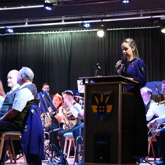 Eine Frau steht an einem Podium und hält ein Mikrofon, während eine Musikband hinter ihr auf der Bühne sitzt. Die Band umfasst verschiedene Instrumente wie Trompeten, Posaunen und ein Schlagzeug. Das Publikum sitzt auf Stühlen, und die Bühne ist mit einem schwarzen Vorhang und Scheinwerfern ausgestattet.