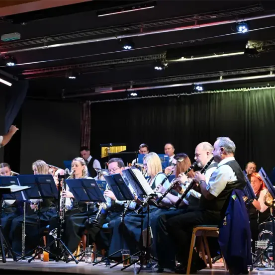 Ein Orchester spielt auf der Bühne mit Musikinstrumenten, darunter Klarinetten und Trompeten, unter Scheinwerfern mit schwarzem Vorhang als Hintergrund.