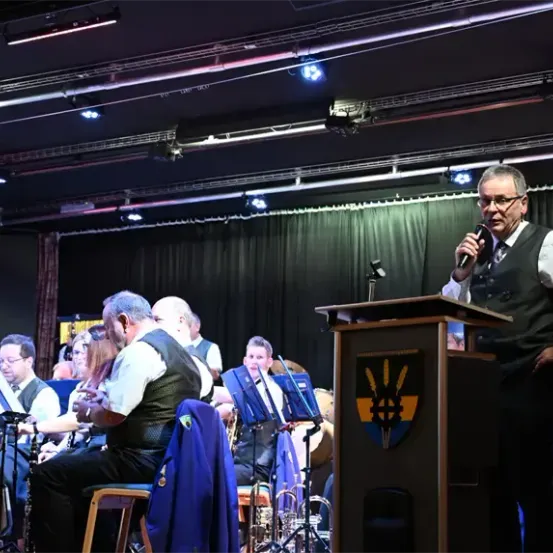 Ein Mann steht an einem Rednerpult mit einem Mikrofon und spricht eine Zuhörerschaft an. Hinter ihm spielt eine Band Instrumente. Scheinwerfer beleuchten die Bühne, und ein Banner hängt im Hintergrund.