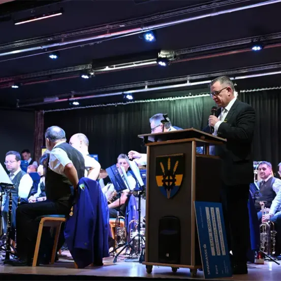 Ein Mann steht an einem Rednerpult auf der Bühne mit einem Mikrofon, möglicherweise spricht er vor einem Publikum. Hinter ihm spielt ein Orchester. Es gibt mehrere sitzende und stehende Personen, einschließlich eines Mannes mit einem blauen Mantel.
