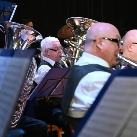Drei Männer in formeller Kleidung spielen Blasinstrumente bei einem Konzert. Der Mann in der Mitte spielt Tuba, während die anderen zwei Trompeten spielen.