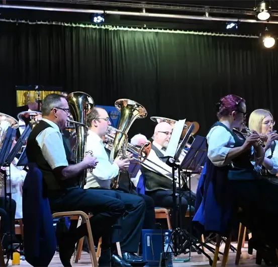 Ein Blasorchester spielt auf der Bühne, Musiker sitzen in Stühlen und spielen Instrumente wie Trompeten, Posaunen und Tuben.