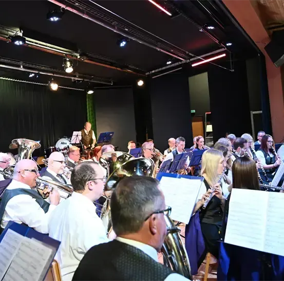 Ein Orchester spielt auf einer Bühne mit verschiedenen Instrumenten. Einige Mitglieder sitzen, andere stehen und halten ihre Instrumente. Die Bühne ist mit roten Lichtern beleuchtet.