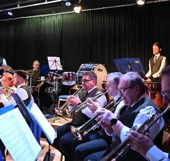 Eine Brassband spielt auf der Bühne, ein Mann in einem Weste spielt hinter ihnen die Trommeln. Die Musiker tragen formelle Kleidung und halten ihre Instrumente. Es gibt auch andere Leute, die auf Stühlen sitzen und Musikinstrumente halten. Es gibt auch einige Notenständer mit Papieren darauf.