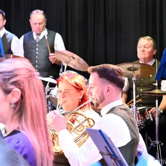 Eine Band spielt auf der Bühne mit verschiedenen Instrumenten. Eine Frau spielt die Trompete, während ein Mann neben ihr lächelt. Dahinter spielen zwei Männer Schlagzeug, und ein weiterer Mann schaut zu.