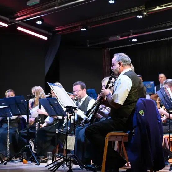Ein Orchester spielt auf der Bühne, wobei Musiker verschiedene Instrumente spielen. Der Mann in der Mitte spielt Klarinette. Andere Mitglieder sitzen und spielen ihre Instrumente. Scheinwerfer sind an der Decke angebracht.