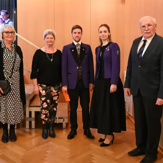 Fünf Personen stehen zusammen, wahrscheinlich bei einer formellen Veranstaltung. Sie tragen formelle Kleidung. Eine Frau in Blumenhosen steht links und ein Mann in einem lila Anzug in der Mitte.