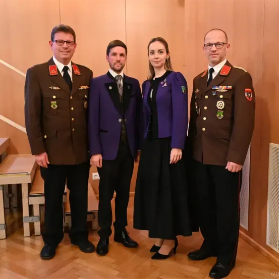 Vier Personen in formeller Kleidung stehen zusammen in einem Raum. Zwei Männer und eine Frau tragen Militäruniformen, während der andere Mann einen Anzug trägt. Sie alle tragen Brillen.