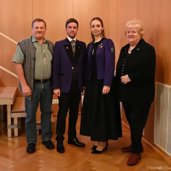 Vier Menschen stehen lächelnd in einem Raum zusammen. Ein Mann in Anzug und eine Frau im Kleid stehen zwischen zwei anderen Frauen. Der Boden ist hölzern, und eine Treppe ist im Hintergrund.