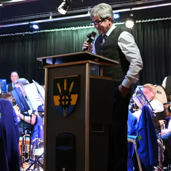 Ein Mann mit Brille steht an einem Rednerpult mit Mikrofon und spricht möglicherweise bei einer Veranstaltung. Hinter ihm spielen Musiker auf der Bühne Instrumente.