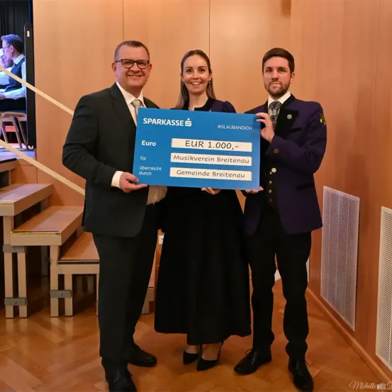 Drei Menschen stehen in einem Raum mit einem blauen Schild, auf dem 'EUR 1.000' steht. Der Mann auf der linken Seite hält das Schild, während die Frau in der Mitte lächelt. Der Mann auf der rechten Seite schaut die Frau an. Hinter ihnen ist eine Holztreppe.