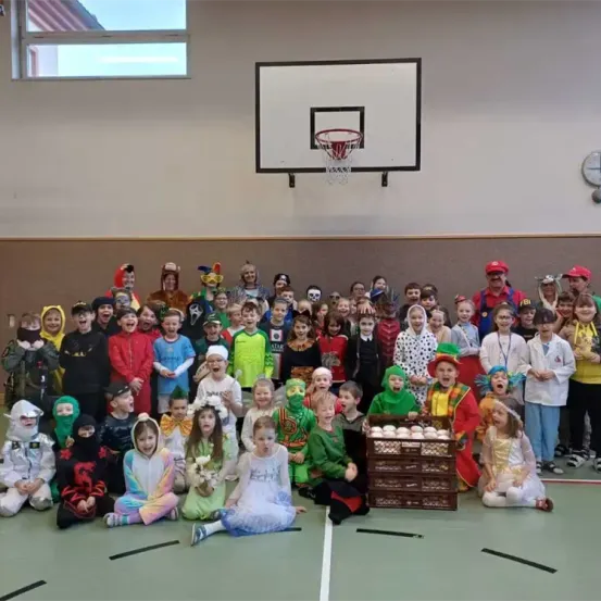 Eine Gruppe von Kindern in Kostümen posiert für ein Foto in einer Turnhalle mit einem Basketballkorb an der Wand.