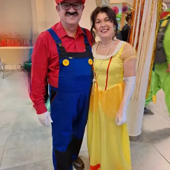 Ein Mann und eine Frau sind in Kostümen gekleidet. Der Mann trägt ein Mario-Kostüm und die Frau ein Schneewittchen-Kostüm. Sie stehen nebeneinander und lächeln.