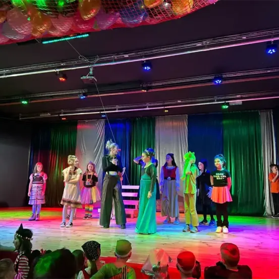 Eine Gruppe von Kindern in Kostümen tritt auf einer Bühne mit bunten Lichtern und einem zuschauenden Publikum auf.