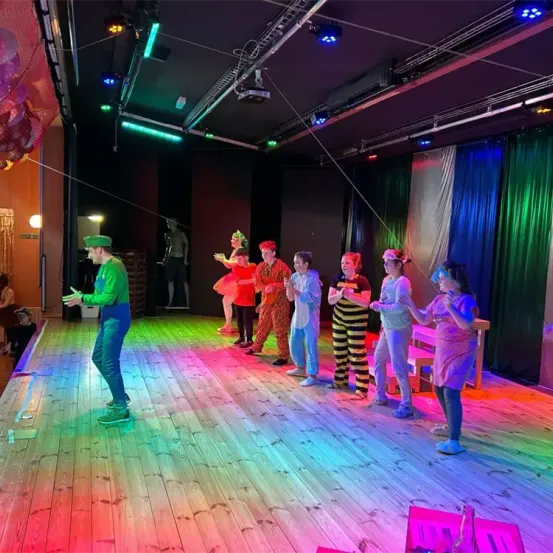 Eine Gruppe von Darstellern in verschiedenen Kostümen steht auf einer farbenfrohen Bühne unter mehrfarbigen Lichtern. Ein Mann gestikuliert mit den Händen.