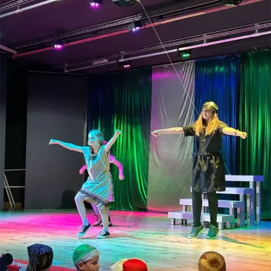 Ein junges Mädchen führt auf einer Bühne mit bunten Lichtern einen Tanz auf. Eine erwachsene Frau steht neben ihr und beobachtet. Das Publikum sitzt vor ihnen.