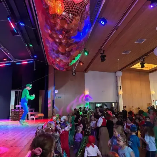 Eine große Gruppe von Kindern in Kostümen versammelt sich auf einer Bühne, mit einem Mann in grüner Mütze und blauer Hose, der auftritt. Bunte Lichter und Luftballons hängen von der Decke.