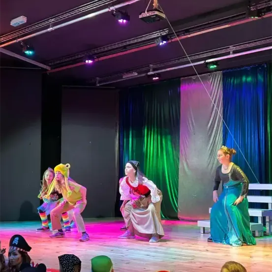 Eine Bühnenaufführung mit vier Frauen, eine in einer gelben Mütze, eine andere in einer roten Mütze und zwei in Kostümen. Dahinter sind farbige Vorhänge.