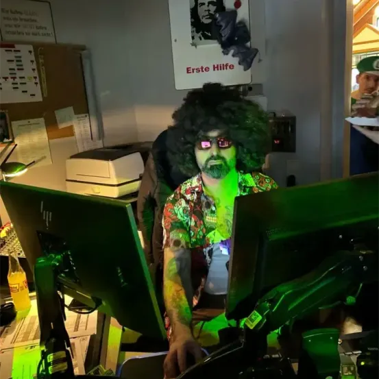Ein Mann mit einer grünen lockigen Perücke und einer Brille sitzt vor einem Computer. Vor ihm stehen zwei Monitore. Dahinter ist ein Plakat mit dem Text 'Erste Hilfe'. Auf dem Schreibtisch steht eine Lampe.