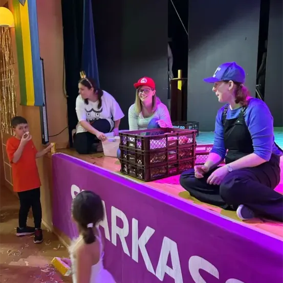 Vier Personen sind auf einer Bühne, möglicherweise während einer Aufführung. Ein Junge isst etwas, ein Mädchen hält ein gelbes Objekt und zwei Frauen sitzen auf der Bühne, eine in einem Hut.