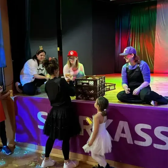 Fünf Personen stehen auf einer Bühne mit einem Banner, das 'SPASS' sagt. Eine Frau hält ein gelbes Objekt, während eine andere kniet. Zwei Frauen sitzen und reden, eine trägt einen roten Hut.