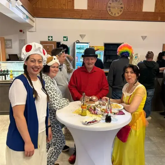Gruppe von Menschen in Kostümen auf einer Party, mit einem Mann in einem roten Hemd und einer Frau in einem gelben Kleid, die an einem Tisch mit Essen sitzen. Eine Uhr und Schilder an der Wand.