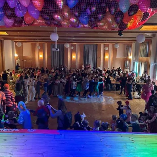 Ein großer Innenraum ist mit bunten Luftballons dekoriert. Eine Menschenmenge, darunter Kinder in Kostümen, tanzt auf dem Boden. Tische und Stühle sind im Hintergrund aufgestellt, und einige Leute stehen herum.