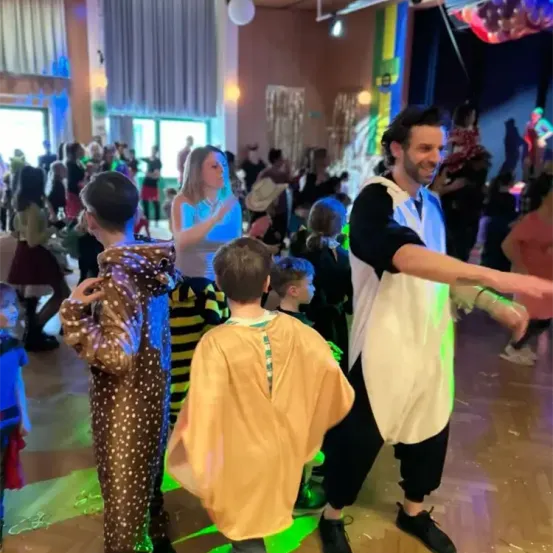 Eine Gruppe von Menschen in Kostümen tanzt in einem Raum. Ein Mann in einem weißen Hemd lächelt und gestikuliert. Hinter ihm schaut eine Frau in einem blauen Kleid zu ihm. Andere Leute tanzen im Hintergrund.